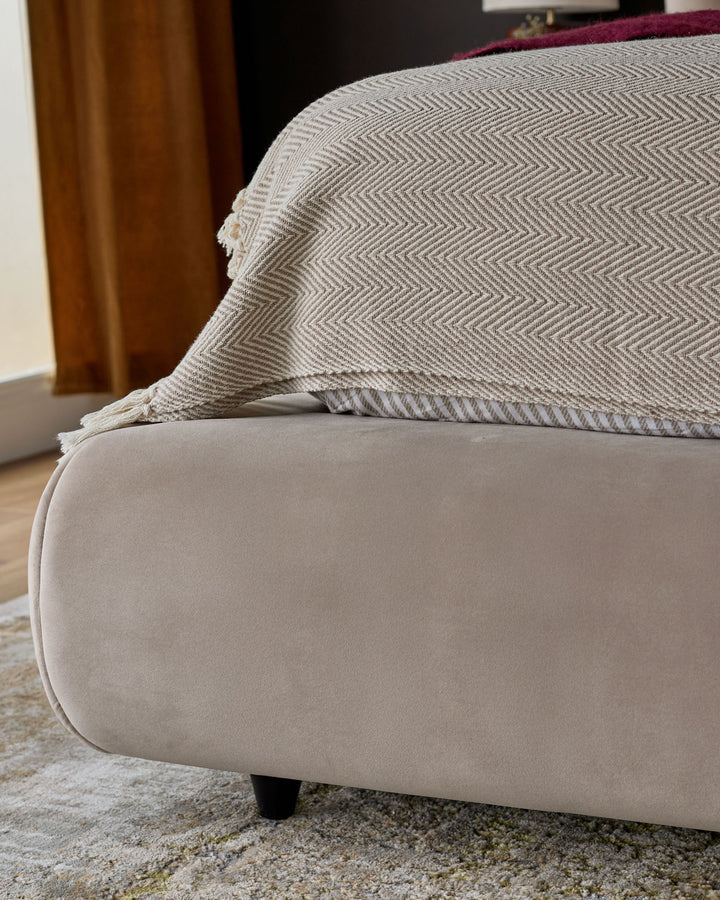 Champagne velvet ottoman bed with herringbone patterned throw blanket, shown from side angle with black legs visible.