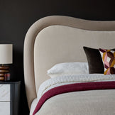 Naomi king-size ottoman bed in champagne velvet with curved headboard and layered bedding, styled with side table and lamp.
