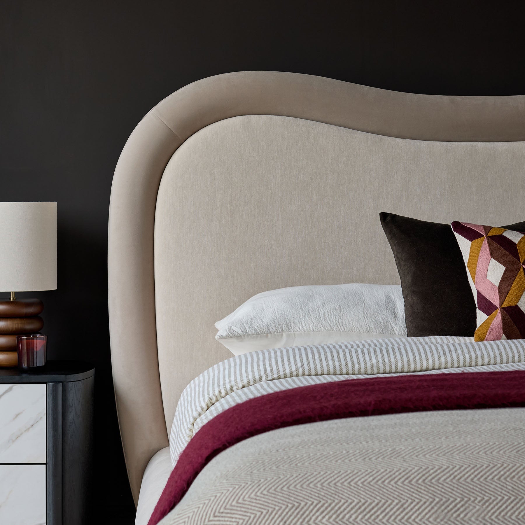 Naomi king-size ottoman bed in champagne velvet with curved headboard and layered bedding, styled with side table and lamp.