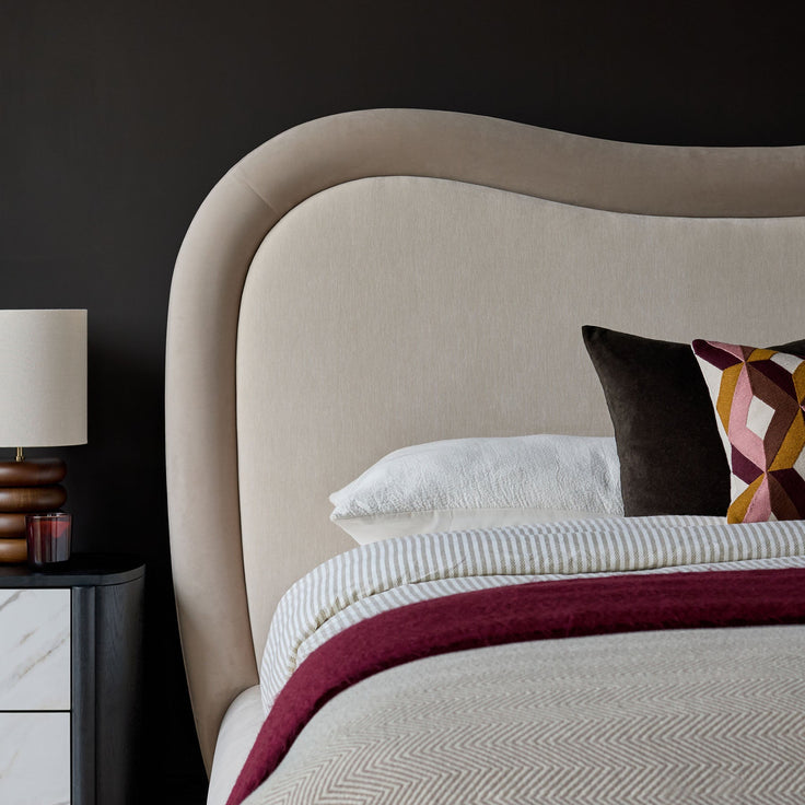 Naomi king-size ottoman bed in champagne velvet with curved headboard and layered bedding, styled with side table and lamp.