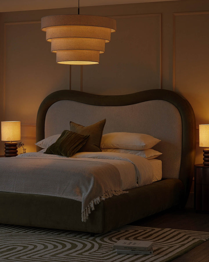 Naomi king-size ottoman bed in olive-green velvet with curved headboard, styled in warm bedroom setting with pendant light and bedside lamps.