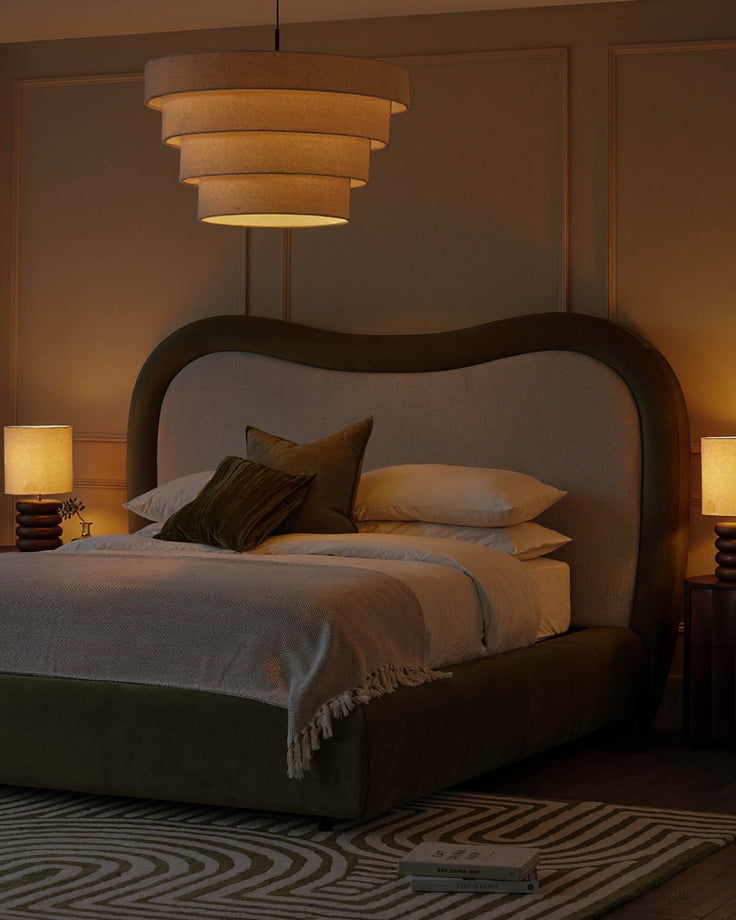 Naomi king-size ottoman bed in olive-green velvet with curved headboard, styled in warm bedroom setting with pendant light and bedside lamps.