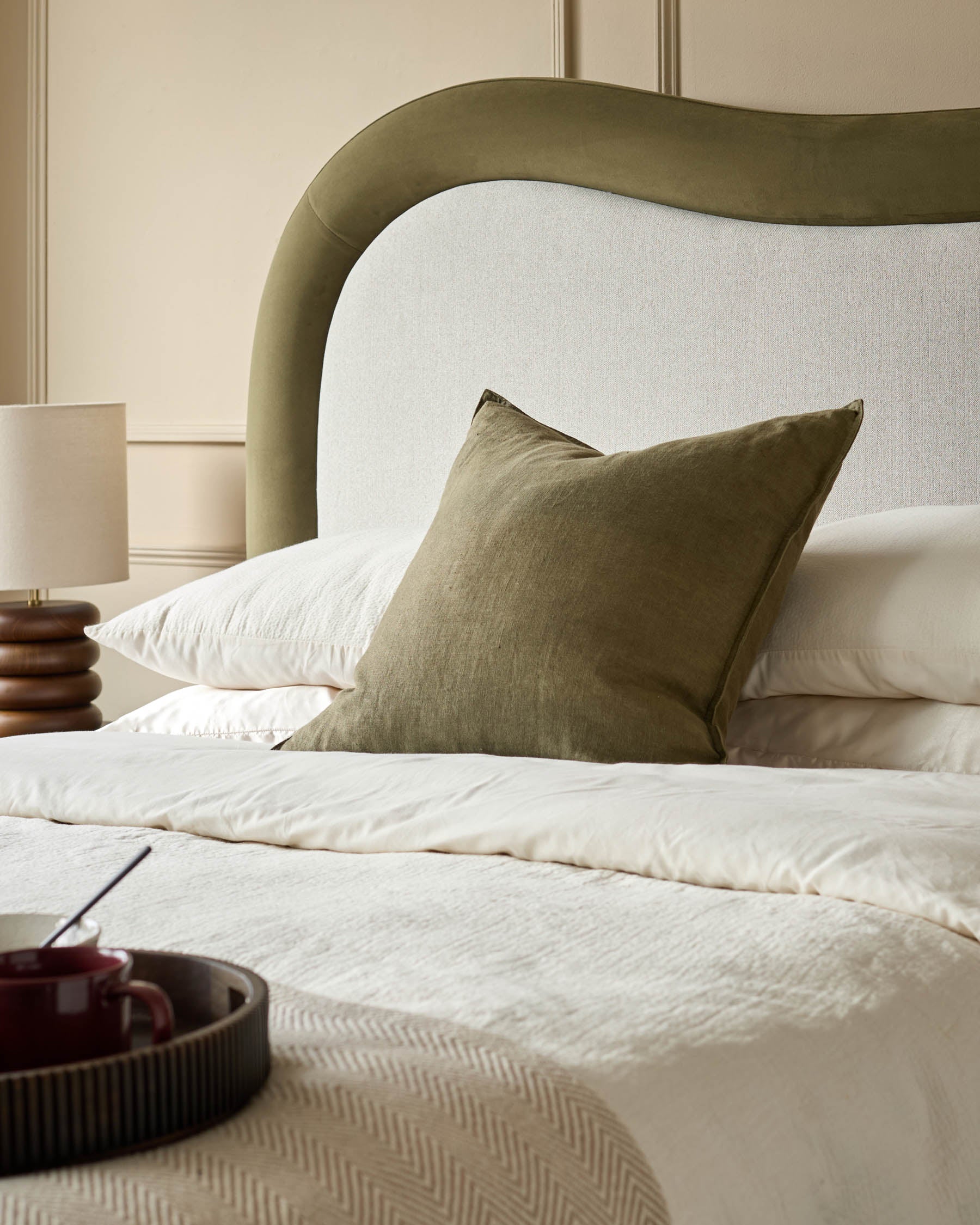 Naomi king-size ottoman bed in olive-green velvet with curved upholstered headboard and cream bedding styled with olive cushion.