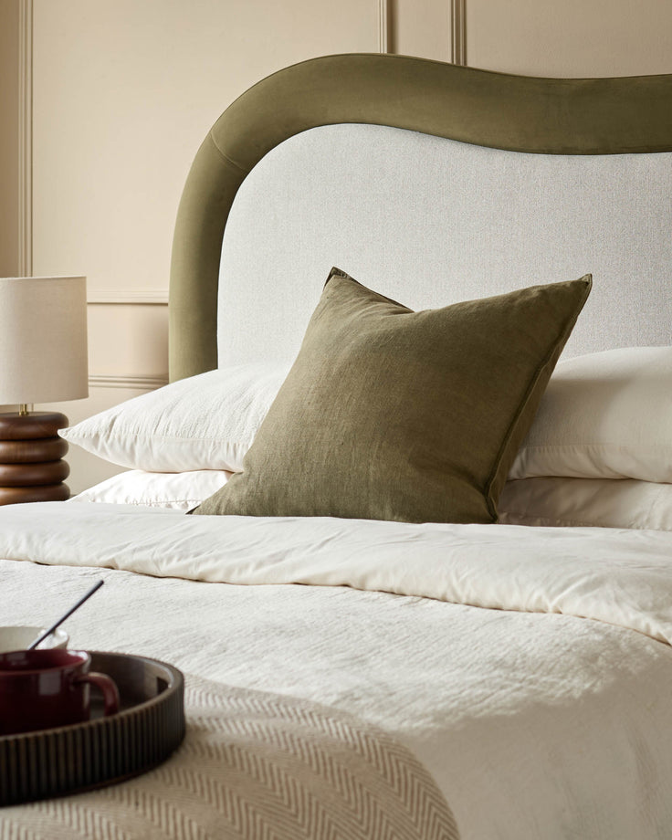 Naomi king-size ottoman bed in olive-green velvet with curved upholstered headboard and cream bedding styled with olive cushion.