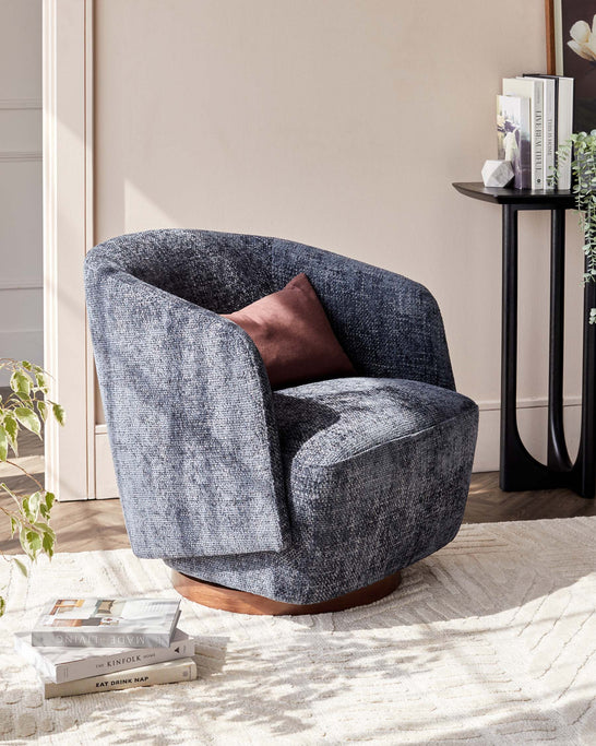 A modern barrel chair upholstered in textured dark blue fabric with a plush seat cushion and a contrasting chocolate brown velvet throw pillow. The chair features a rounded back and a unique circular wooden base, set against a neutral interior with a black side table, decorative books, and a potted plant, all atop a cream textured area rug.