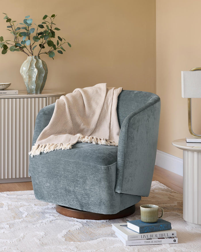 A modern, teal swivel armchair with a beige throw, styled with a side table and books on a patterned rug in a warm, neutral room.