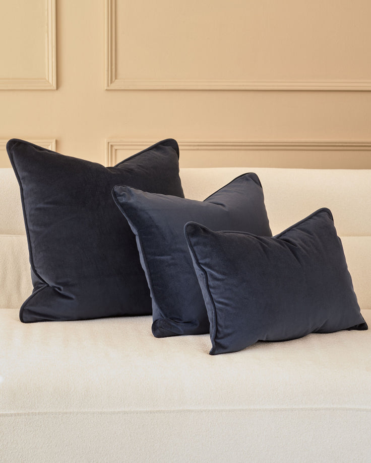 Navy velvet cushion in medium size displayed on a cream sofa against a paneled wall backdrop.