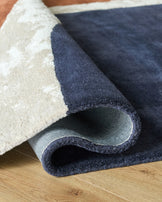 Close up of a plush, patterned rug in neutral and blue tones, partially rolled on a light wooden floor.