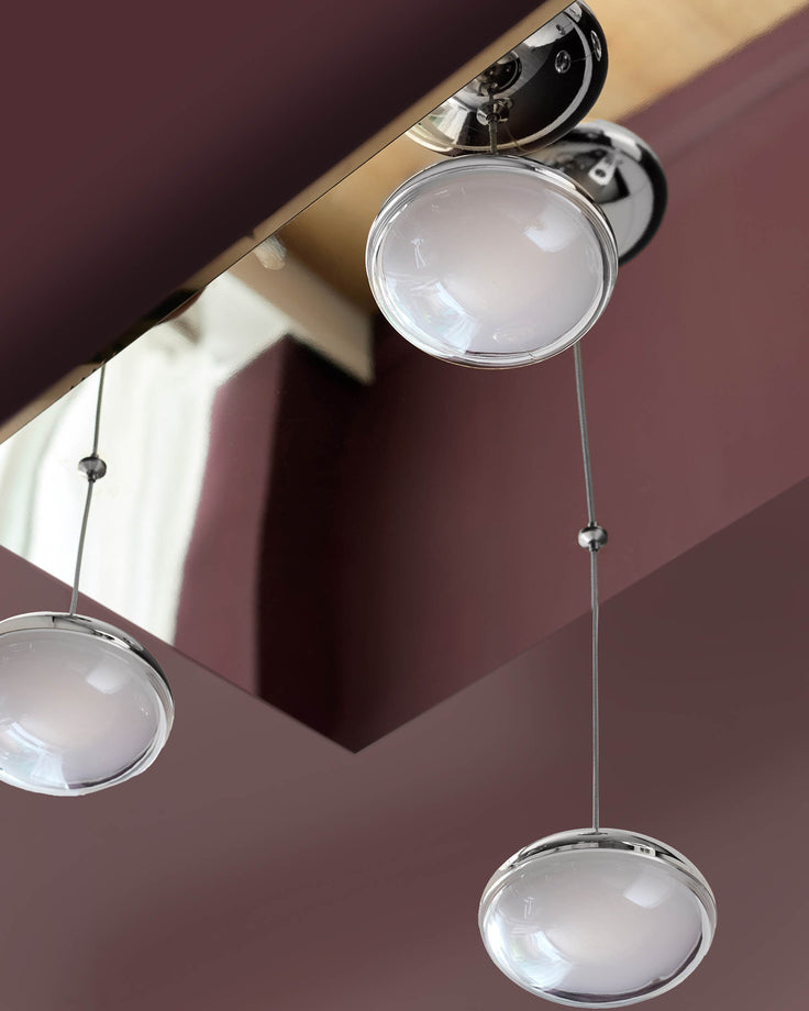 Nina 7-Pendant Light with clear glass domes and chrome hardware suspended from a wooden ceiling beam against a mauve wall.