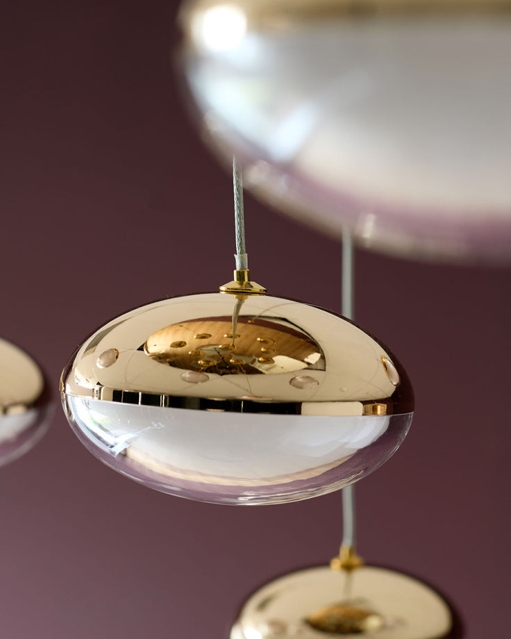 Nina pendant light with clear glass dome and soft gold metal fixture, hanging against mauve background.