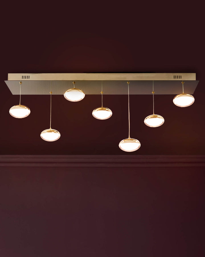 Nina 7-Pendant Light in soft gold with round frosted glass shades suspended from a rectangular metal canopy against a dark wall.