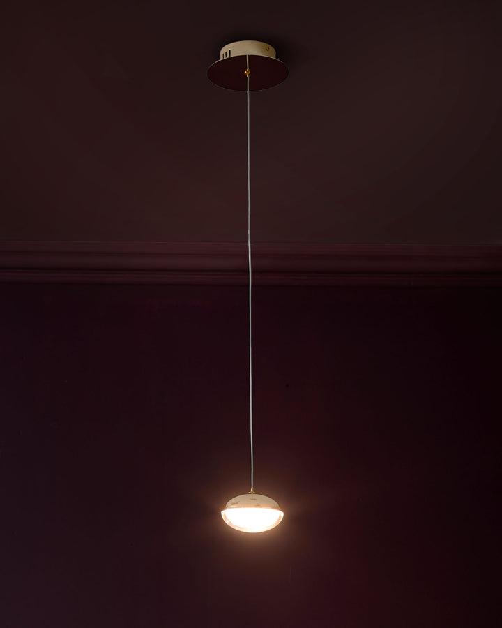 Nina single pendant light with soft gold finish and frosted glass dome shade, hanging from thin cord against dark wall.