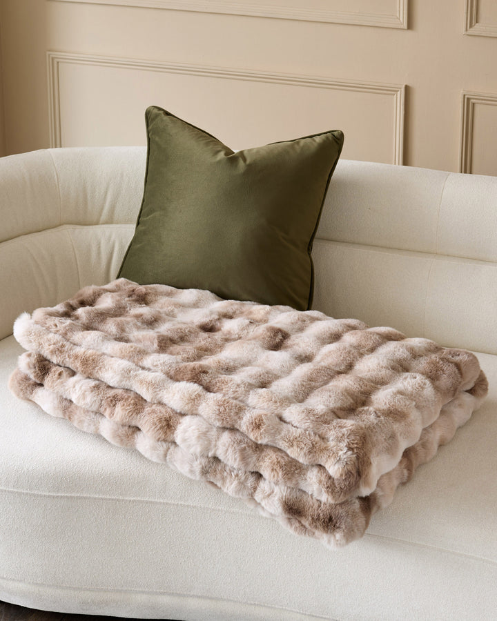 Nova throw blanket in natural beige, quilted texture, draped on cream curved sofa with olive green pillow.