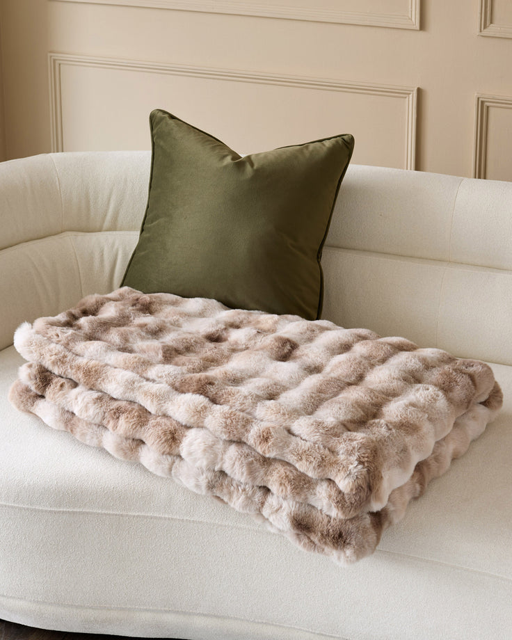 Nova throw blanket in natural beige, quilted texture, draped on cream curved sofa with olive green pillow.