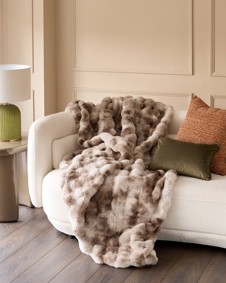 Nova throw blanket in natural, draped over a cream curved armchair with decorative pillows in a neutral living room setting.