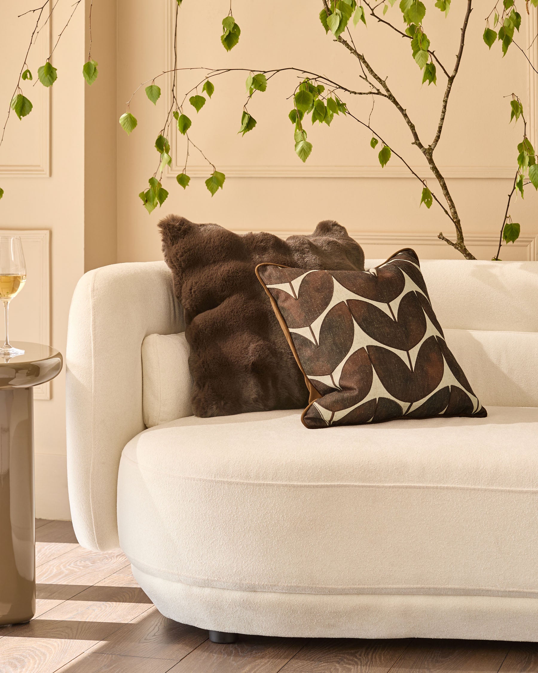 Chocolate brown cushion with geometric leaf pattern, displayed on cream curved sofa in styled living room setting.