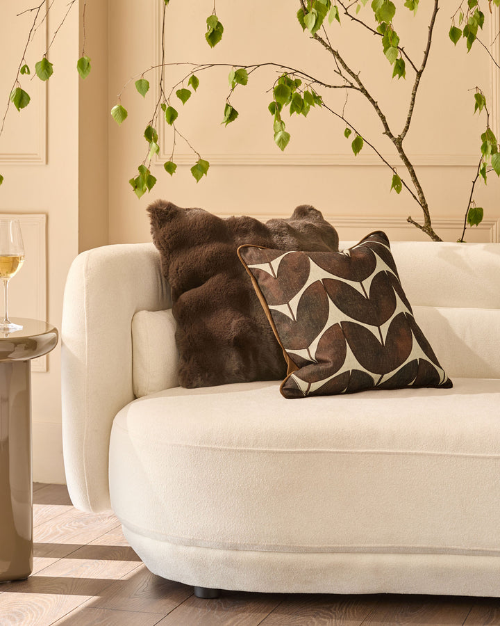 Chocolate brown cushion with geometric leaf pattern, displayed on cream curved sofa in styled living room setting.