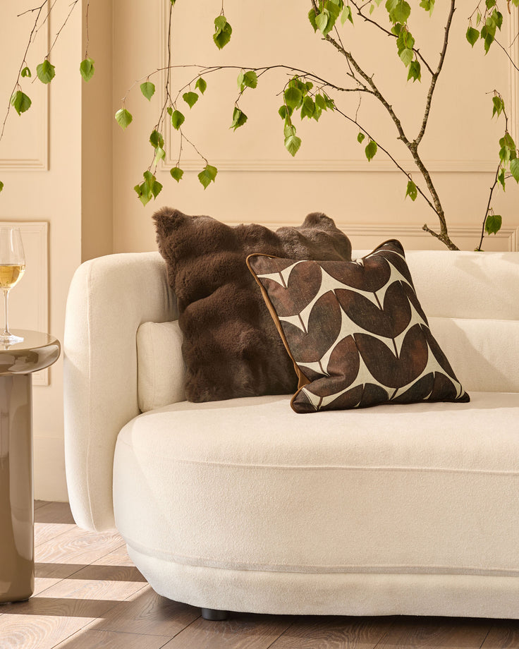 Chocolate brown cushion with geometric leaf pattern, displayed on cream curved sofa in styled living room setting.