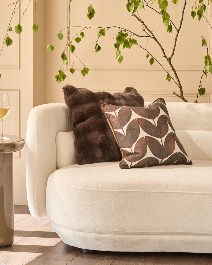 Large chocolate cushion with geometric leaf pattern, displayed on cream curved sofa in modern living room setting.