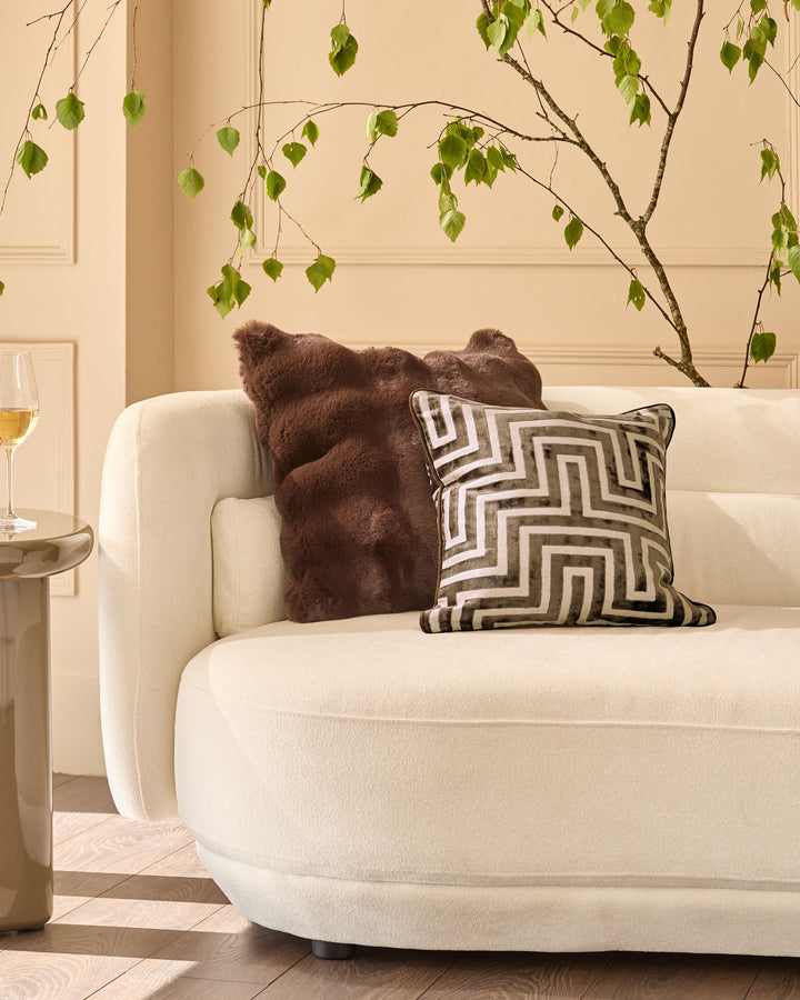 Chocolate brown faux fur cushion displayed on a cream armchair with geometric patterned pillow nearby.