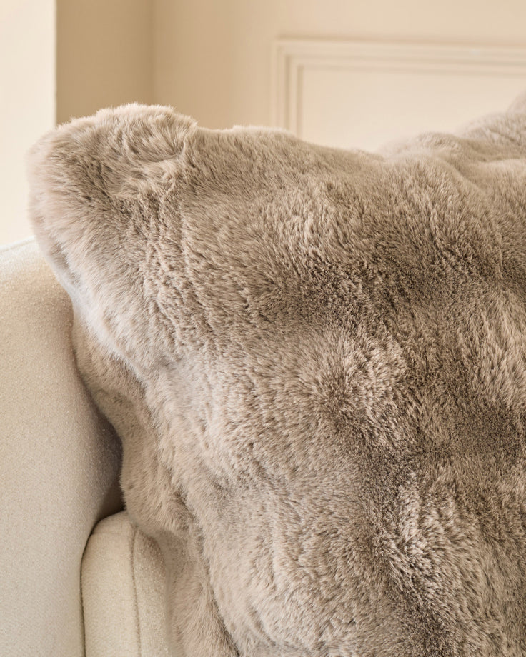 Large taupe cushion with plush texture, displayed on a cream upholstered chair in a bright interior setting.