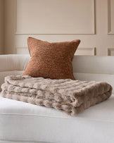 Nova throw in taupe, draped over a cream sofa with a rust-textured pillow against a paneled wall.
