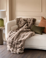 Nova Throw Large in Taupe draped over a cream curved sofa with coordinating pillows in a neutral bedroom setting.