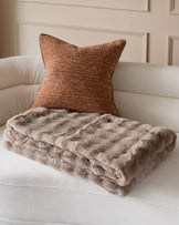 Nova throw in taupe draped over cream upholstered chair with rust textured pillow, showing quilted texture detail.