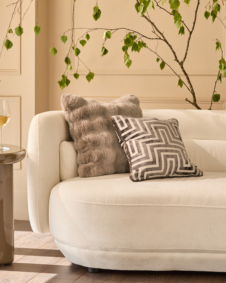 Large taupe cushion with textured knit pattern, displayed on cream curved sofa in modern living room setting.