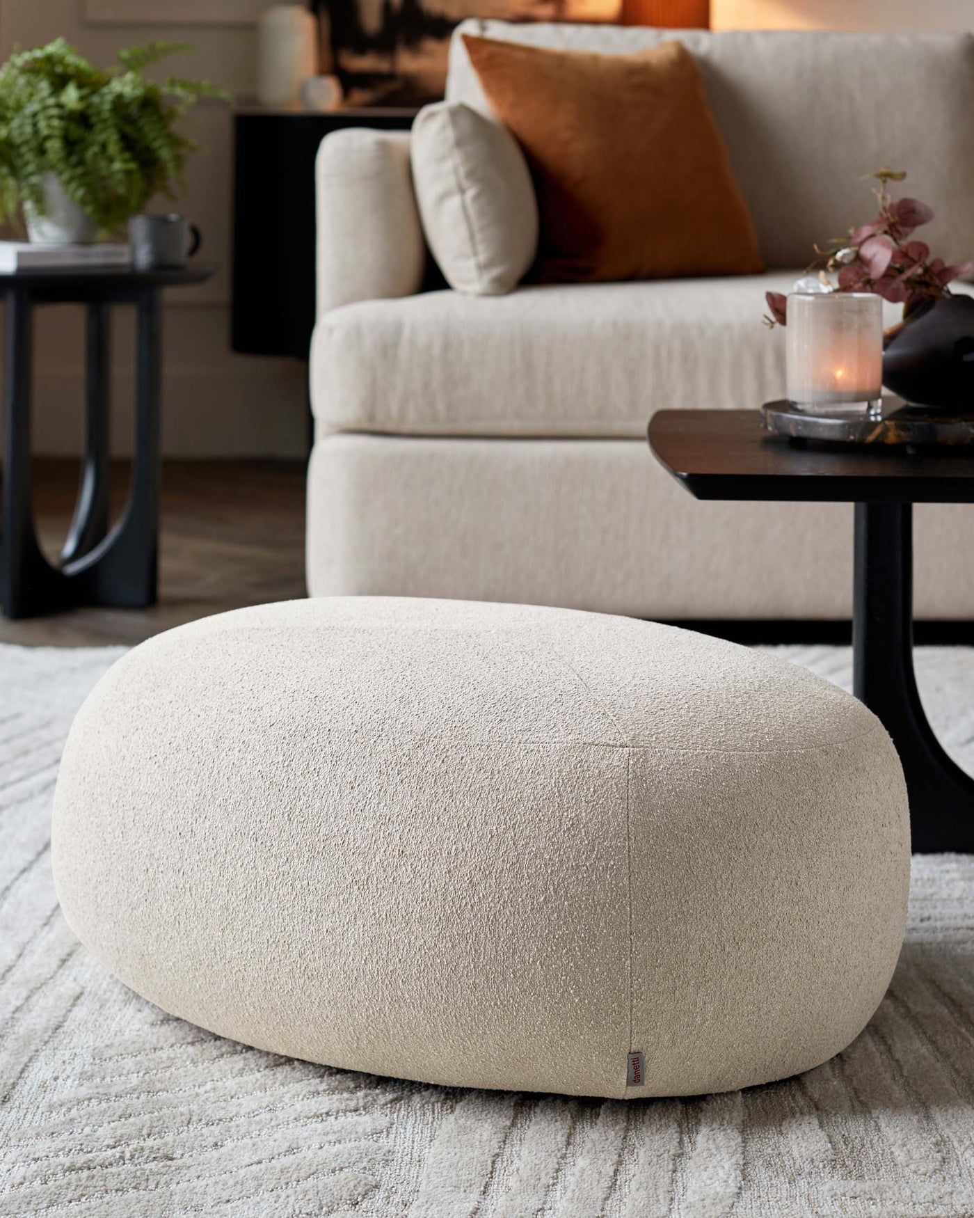Elegant living room featuring a contemporary beige sofa with plush cushions and a round beige boucle ottoman in the foreground. A small round black side table with a candle is visible next to the sofa, all resting on a textured light area rug.