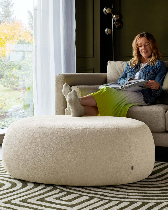 Beige upholstered oval ottoman with a textured fabric, positioned on a patterned area rug in a cosy living space setting.