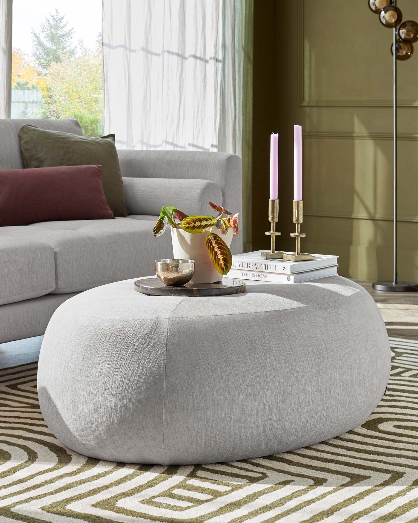 Contemporary living room with a light grey upholstered round ottoman at the centre. The ottoman is complemented by a grey fabric sofa adorned with neutral-toned cushions, resting against a window with sheer curtains. A geometric pattern rug in cream and olive tones underlies the furniture setup. Decorative elements include a plant, candles, and books atop the ottoman.