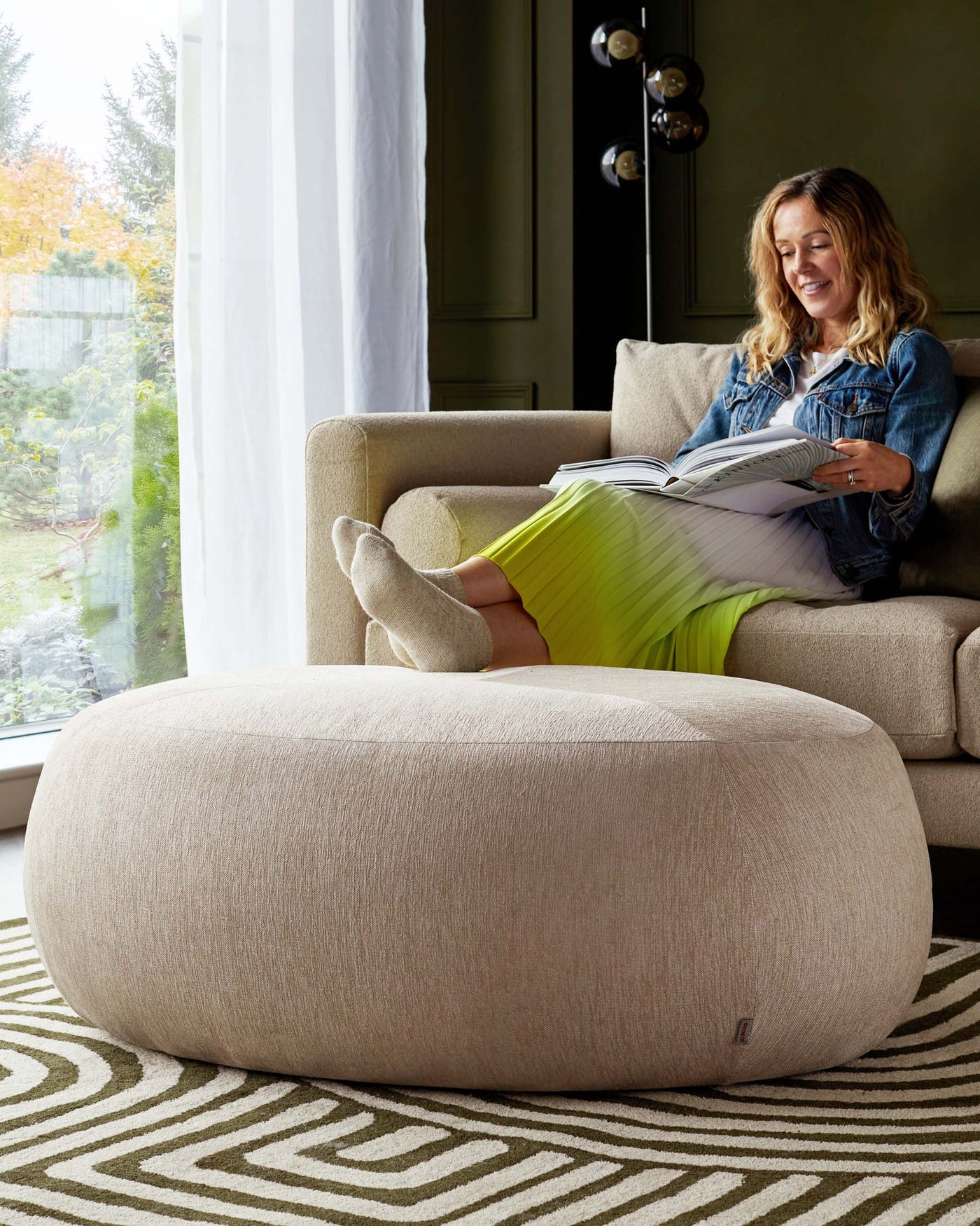 A contemporary beige fabric upholstered ottoman with a cylindrical silhouette in front of a coordinating beige armchair, set upon a patterned area rug with geometric lines.