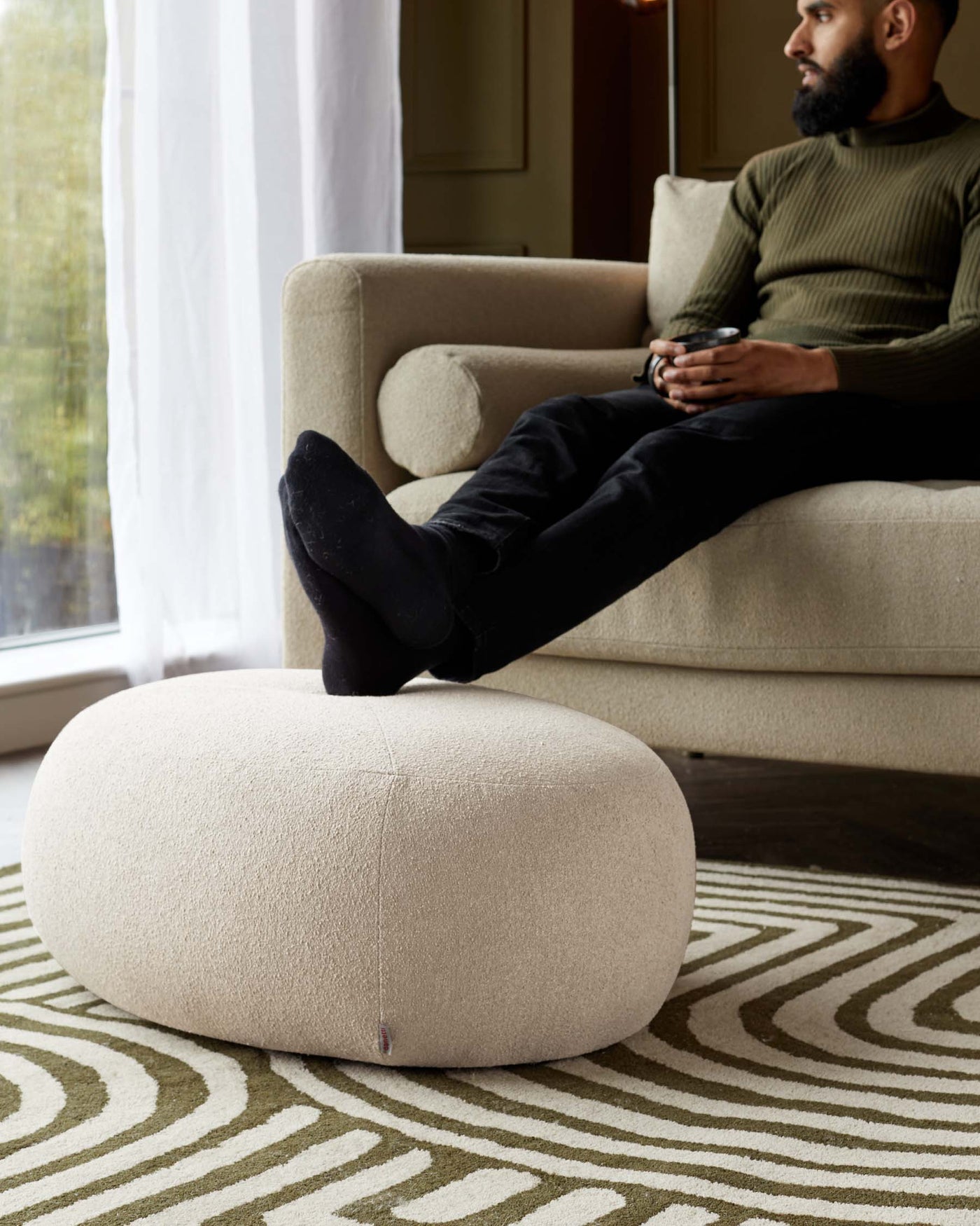Beige upholstered sofa with a matching plump round ottoman on a patterned area rug.