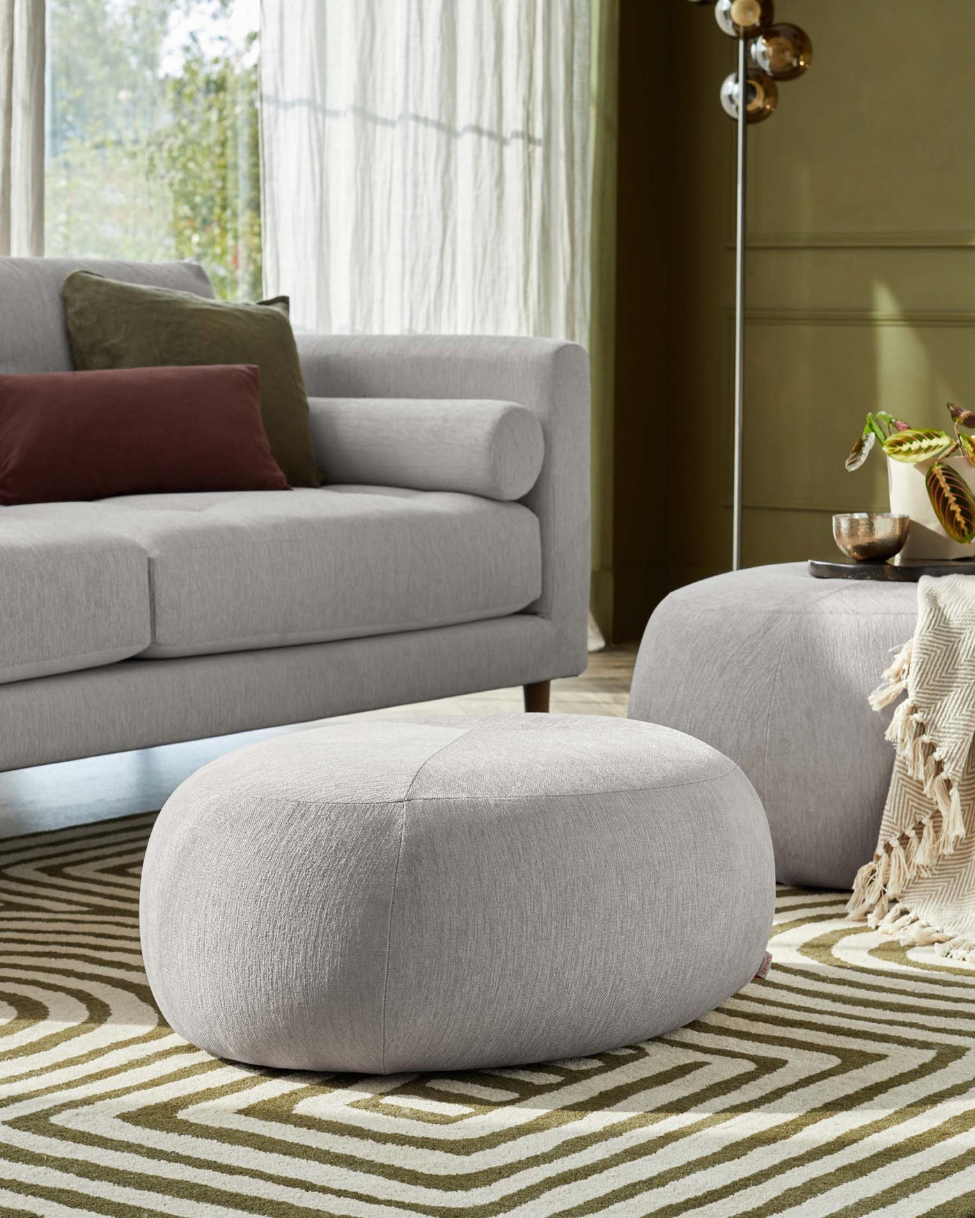 A modern light grey upholstered sofa with clean lines and cylindrical cushions on the sides. In front of the sofa are two round grey fabric ottomans with a subtle textural design. The furniture is displayed in a room with natural light, a geometric-patterned rug on the floor, and a hint of a metallic floor lamp in the background.
