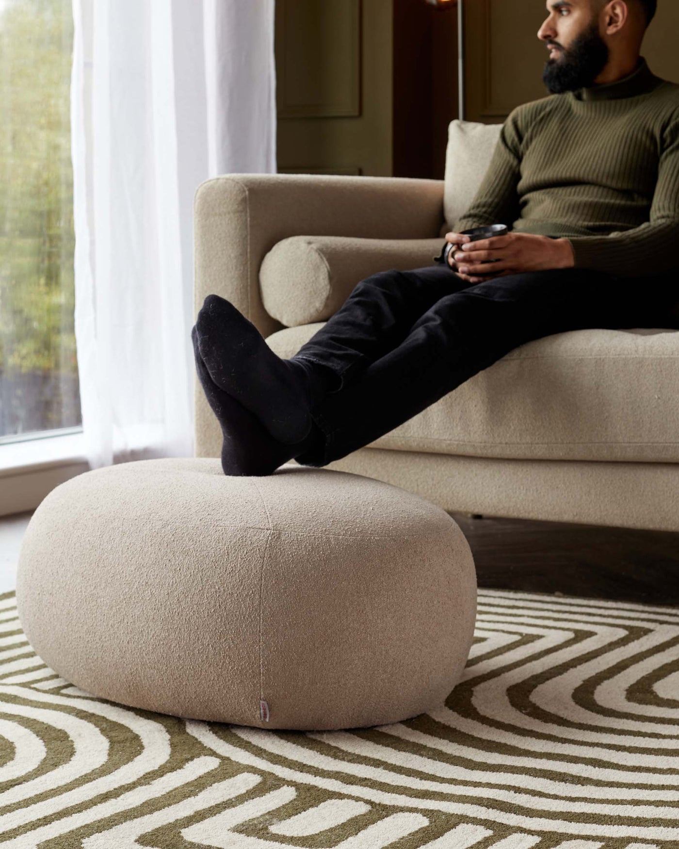 A modern beige fabric upholstered armchair with a matching circular ottoman on a patterned area rug. The armchair features a clean-lined contemporary design with plush cushioning, rounded armrests, and a snug backrest. The ottoman is a simple cylindrical shape, providing a comfortable footrest or additional seating option. Both pieces exhibit a minimalist aesthetic with a focus on comfort and style.