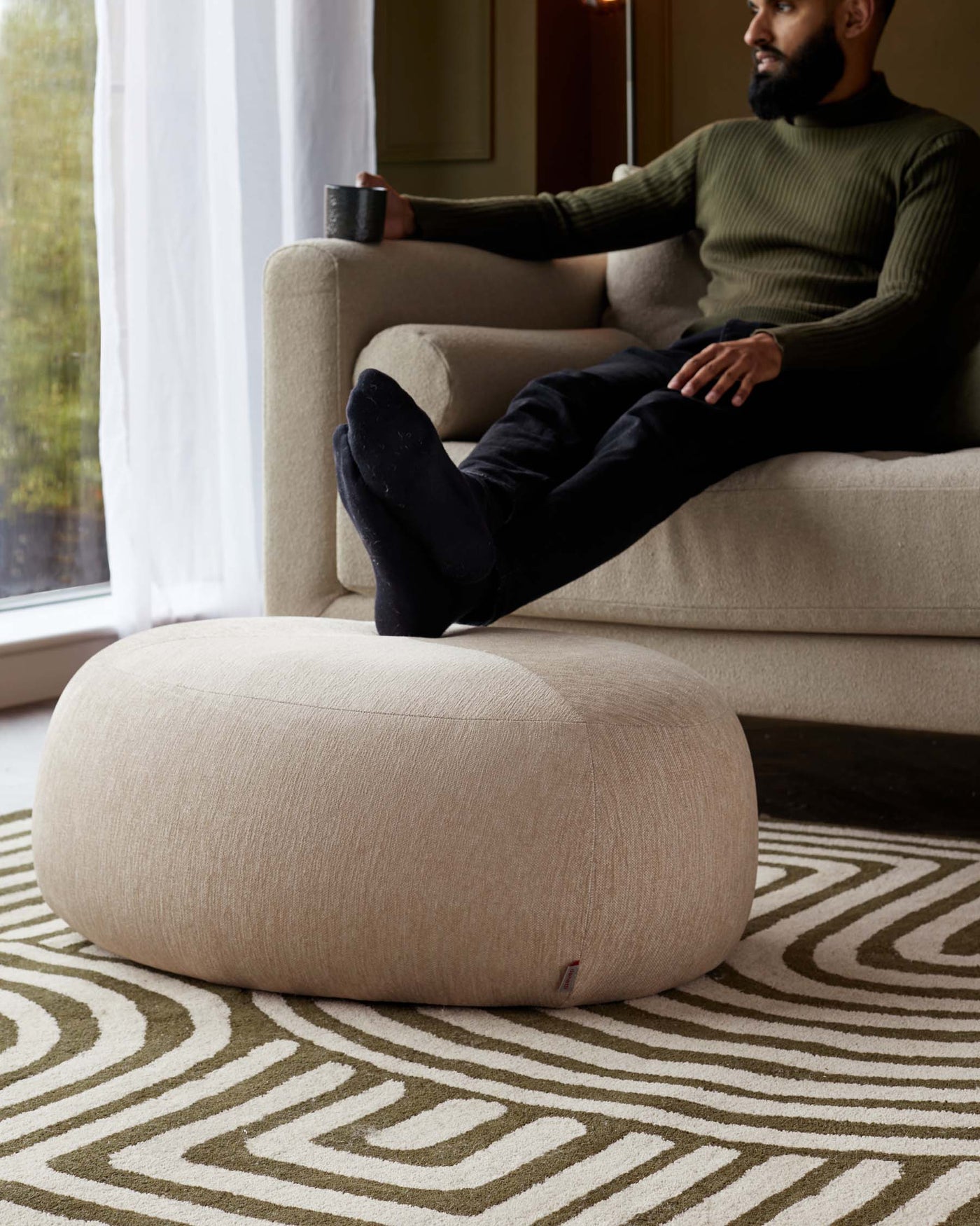 A contemporary beige fabric sofa with a clean, minimalist design, complemented by a coordinating round beige fabric ottoman, set against a geometric patterned rug in earth tones.