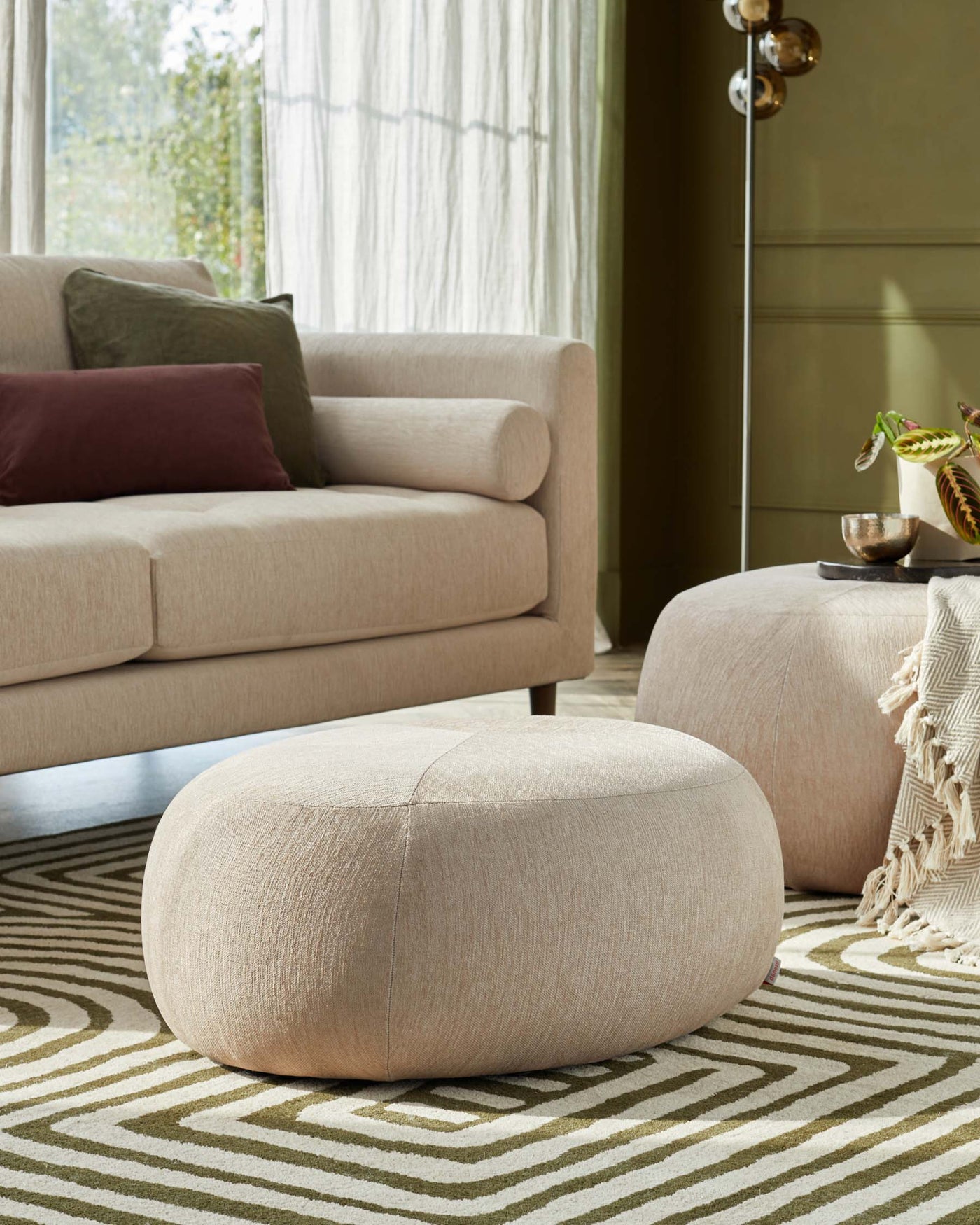 Contemporary beige sofa with plush cushions and clean lines, complemented by two round, fabric-upholstered ottomans in a matching hue, all set upon a geometric-patterned area rug in earth tones.