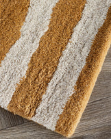Small sculpted wool rug with abstract swirl pattern in ochre and cream, shown at an angle on a wood floor.