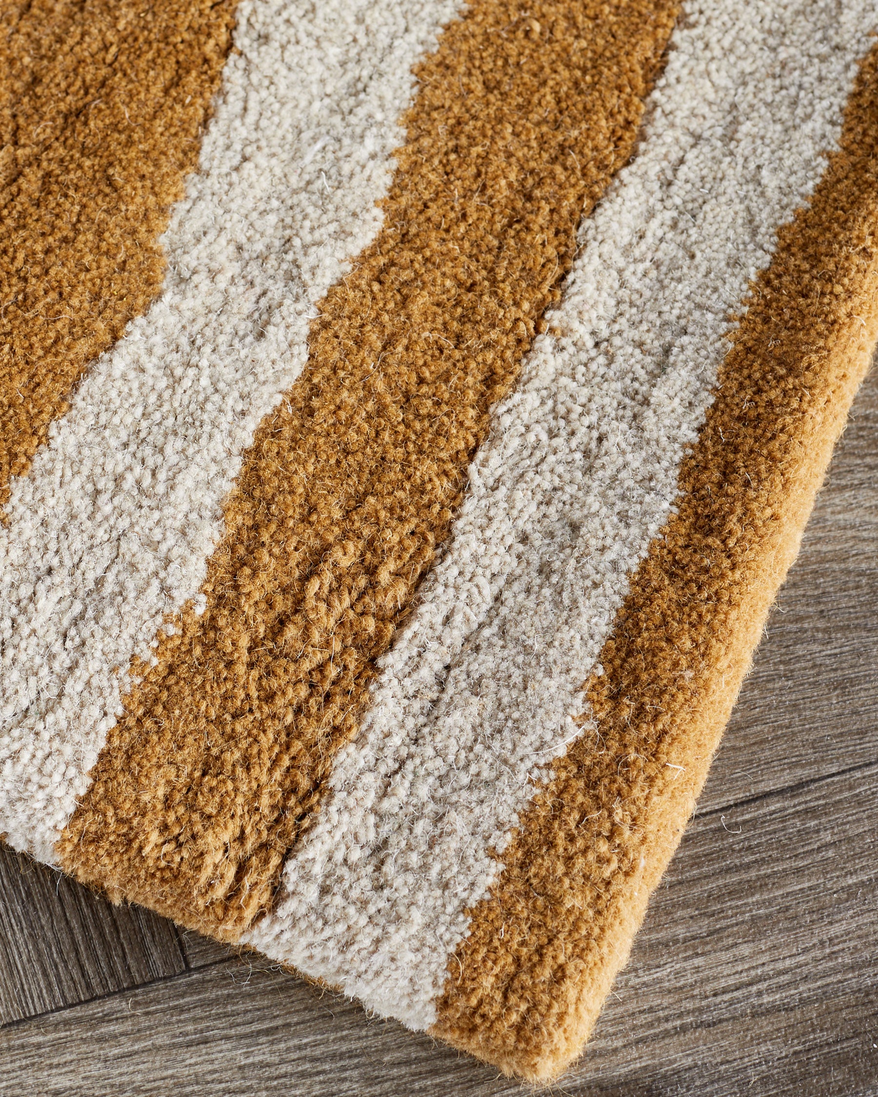 Small sculpted wool rug with abstract swirl pattern in ochre and cream, shown at an angle on a wood floor.