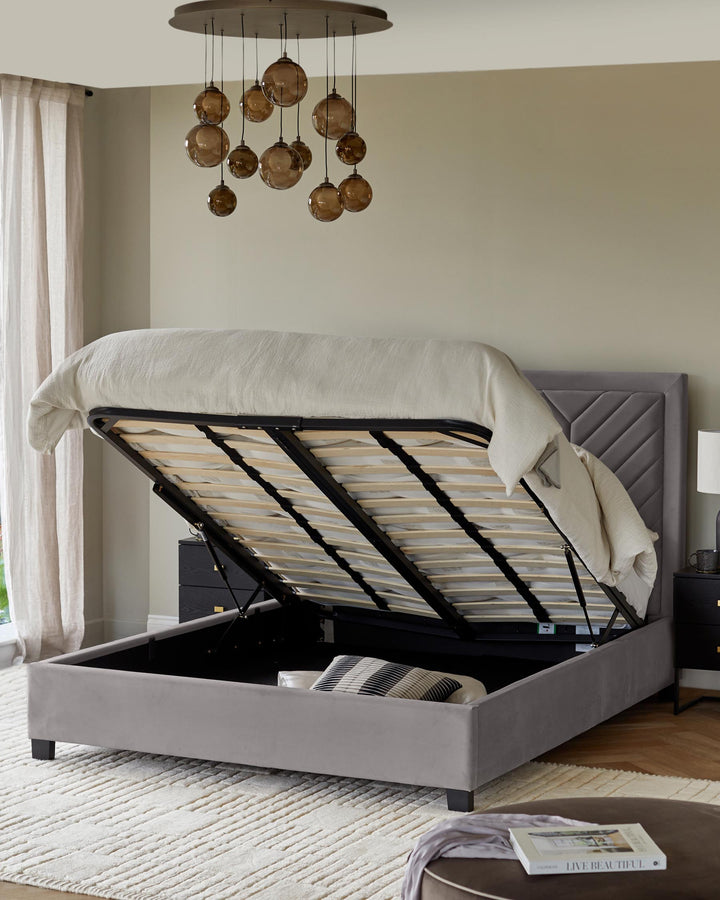 Grey velvet ottoman storage bed with a headboard, brown hanging lights, side table, and chest of drawers.