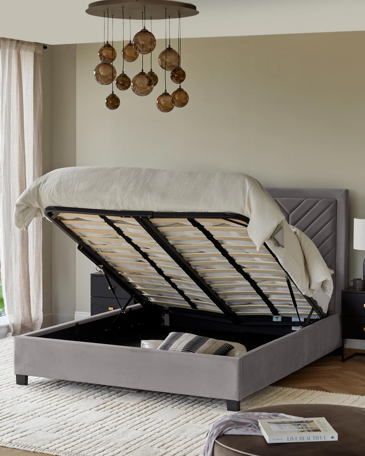 Grey velvet ottoman storage bed with a headboard, brown hanging lights, side table, and chest of drawers.