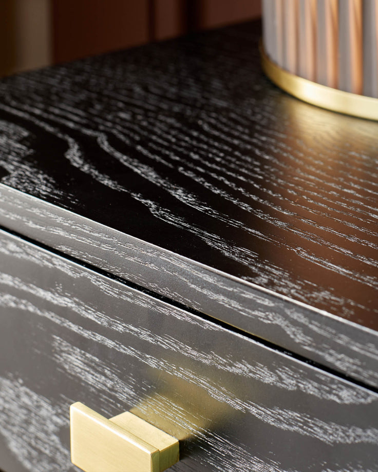 Close-up of a black wood grain bedside table, featuring a brass drawer handle and stylish lamp.