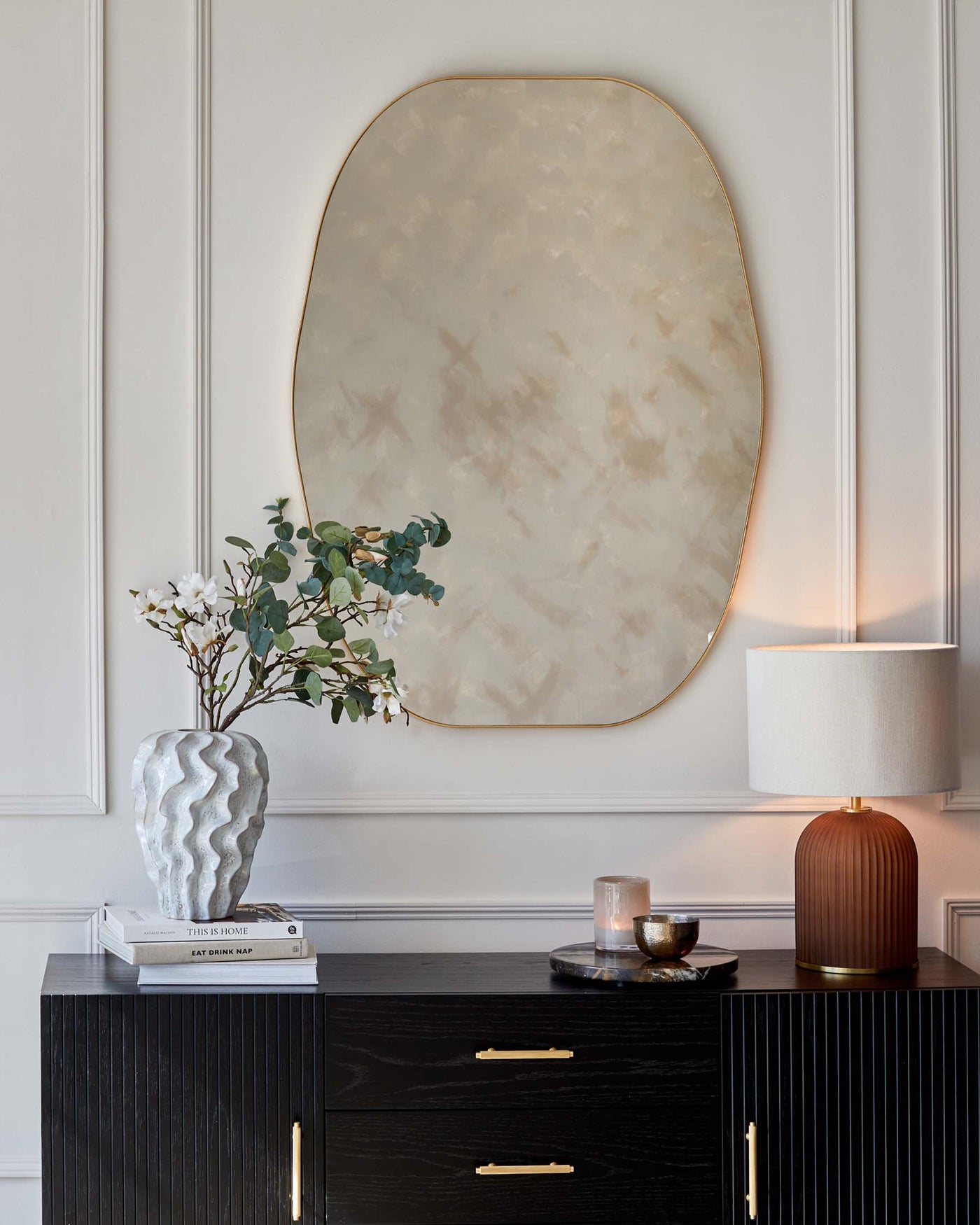 Elegant contemporary black sideboard with brass handles, set against a detailed white panel wall, complemented by a decorative wavy white vase on a stack of hardcover books, with a cylindrical lamp featuring a wood-textured base and white shade on one end of the sideboard, next to a tray holding a candle and cup. A large, abstract cloudy mirror with a subtle gold frame hangs above, anchoring the sophisticated vignette.