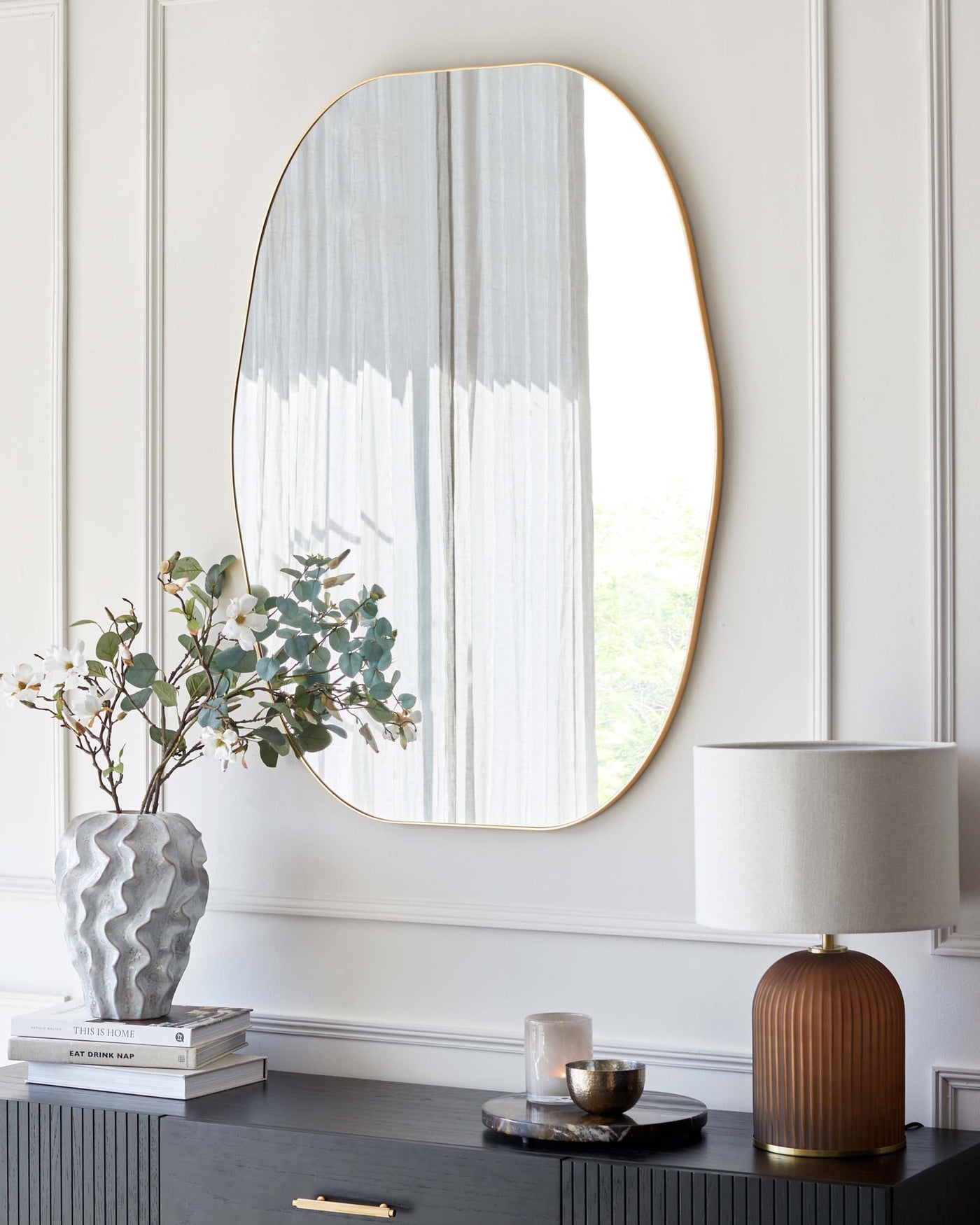 A sleek modern room featuring an elegant oval mirror with a thin gold frame mounted on a white wall with decorative mouldings. Below the mirror is a dark wood console table with ribbed frontal detailing and gold handles. The table is adorned with a tall, textured grey vase holding eucalyptus branches, a stack of hardcover books, a cylindrical white lamp with a brown base, and a small round dish with a candle. A round black side stool with a glossy finish sits adjacent to the console table.