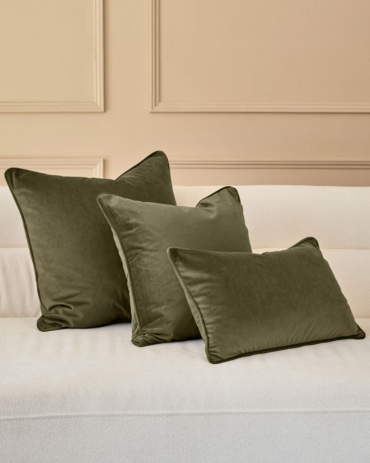 Luca medium cushion in olive green velvet, displayed on a cream bed against a beige paneled wall.