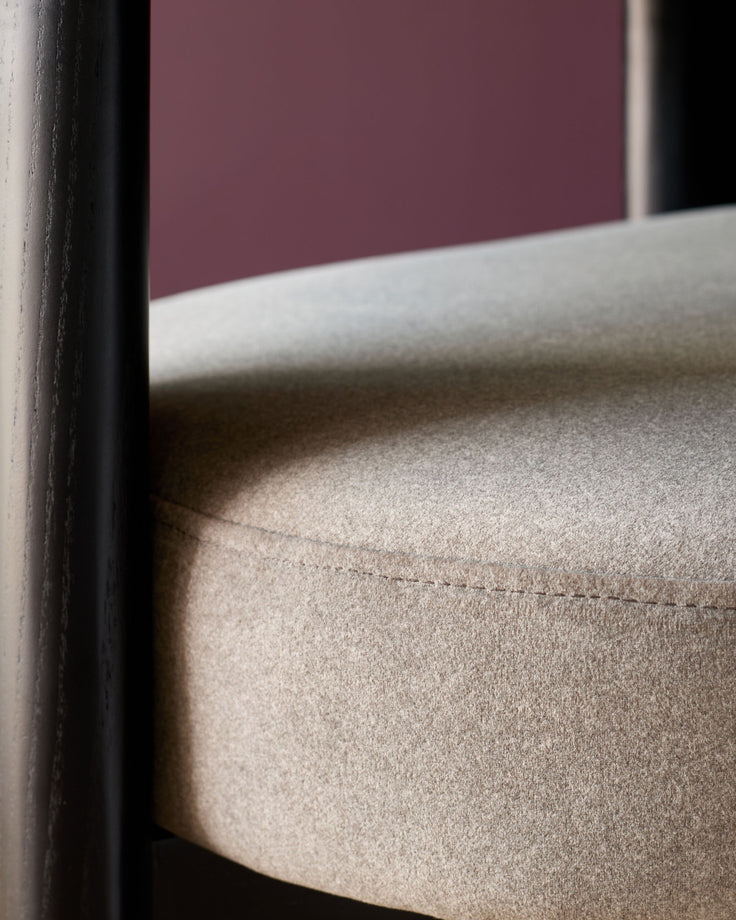 Close-up of a chair seat with a tan fabric cover and dark wooden frame. The seat is rounded with a visible seam detail.