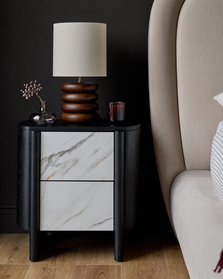 Orlan bedside table in black wood with white marble ceramic drawers, styled with wooden lamp and decor.
