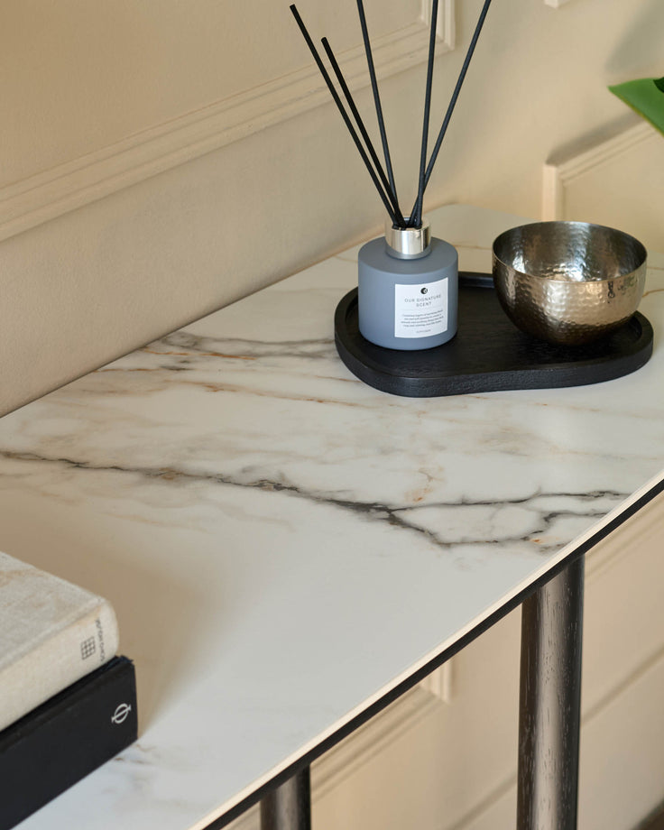 A modern console table with a faux marble top and dark metal legs. It is styled with a diffuser, bowl, and books.