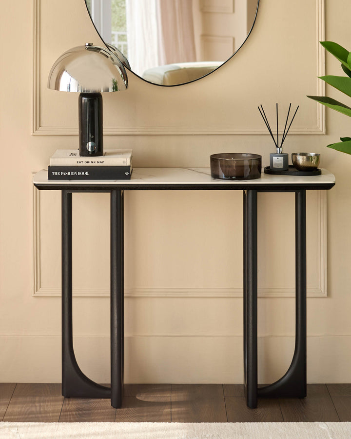 A console table with marble top and black legs displaying books, a lamp and fragrance diffusers.